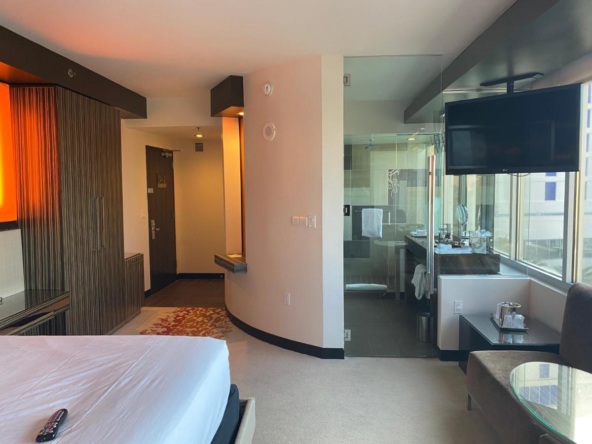 Hard Rock Hotel & Casino Biloxi Exterior foto The photo shows a modern hotel room. On the left side, there is a bed with white bedding, and the headboard area features warm lighting. The room has a window that allows natural light to come in, offering a view of the outside. On the right side, th